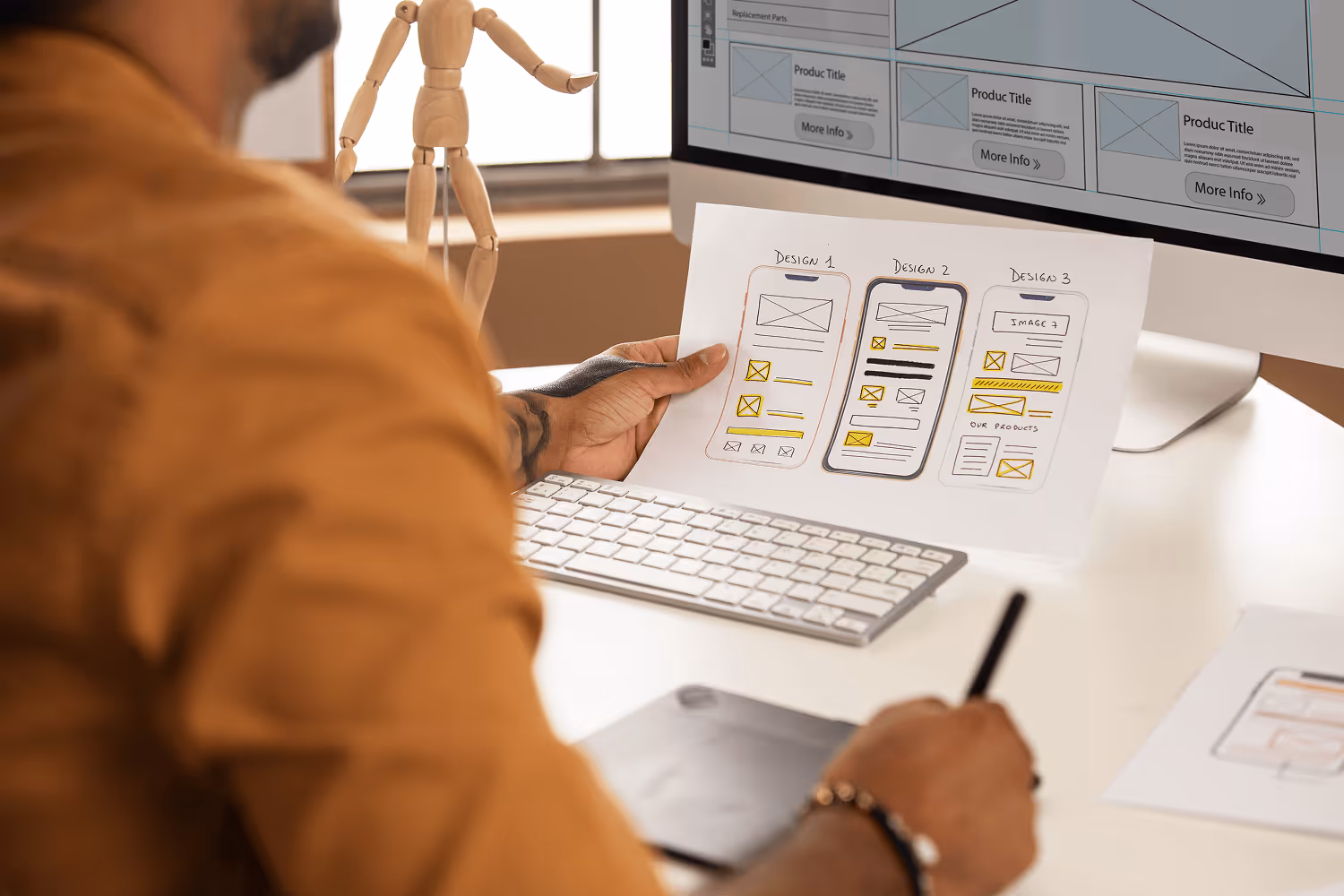 Designer holding paper with three mobile app wireframe sketches in front of a computer screen with website mockup.