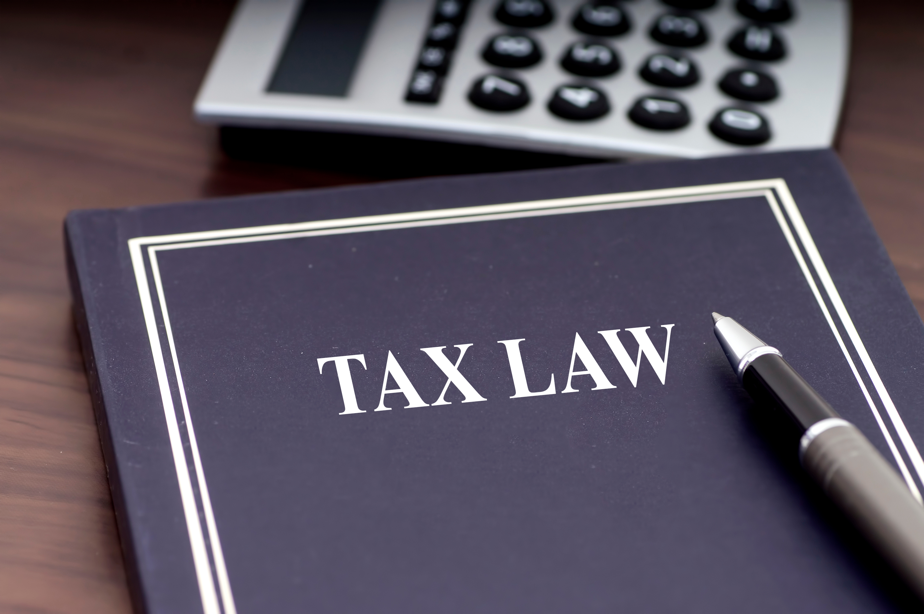 Close-up of a 'TAX LAW' book on wood surface with pen and calculator, symbolizing legal expertise and financial analysis.