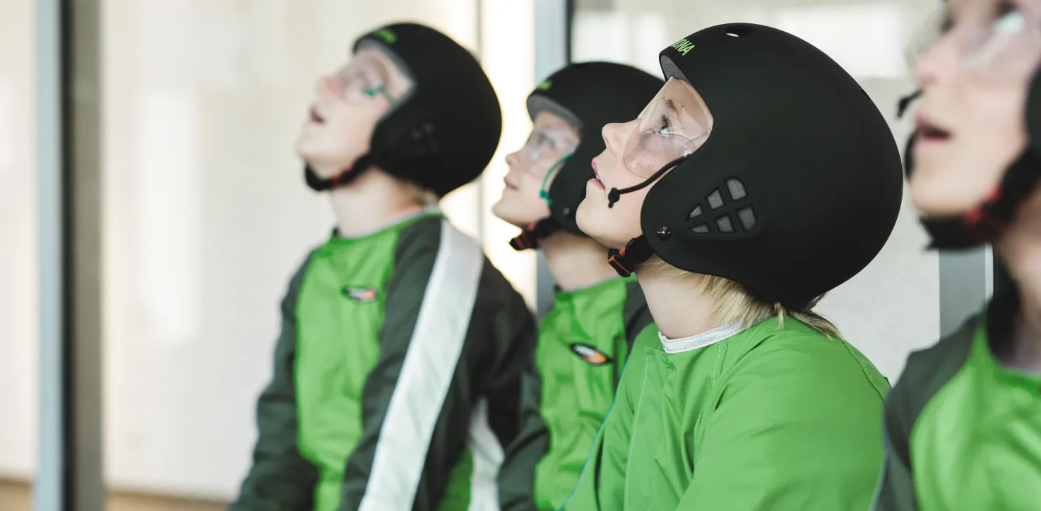 Children in flight suits and helmets watching skydiving session.