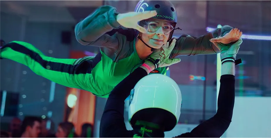 Person experiencing indoor skydiving in a vertical wind tunnel, supported by an instructor wearing a helmet.