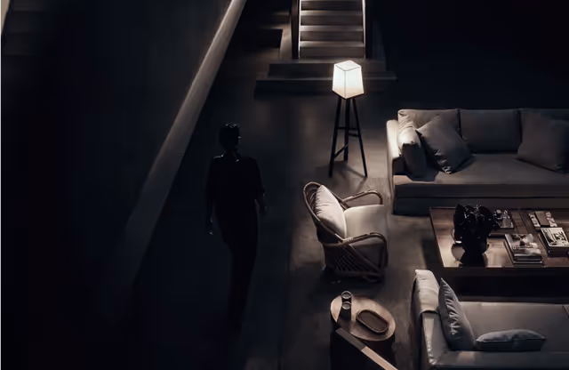 Dimly lit modern living room with a person walking, featuring sofas, armchair, coffee table, and a standing lamp near stairs.