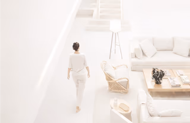 Woman dressed in white walking barefoot in a bright, minimalist living room with light-coloured sofas, a wooden coffee table, and a wicker chair.