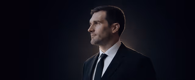 Profile portrait of a man in a black suit and tie against a dark background.