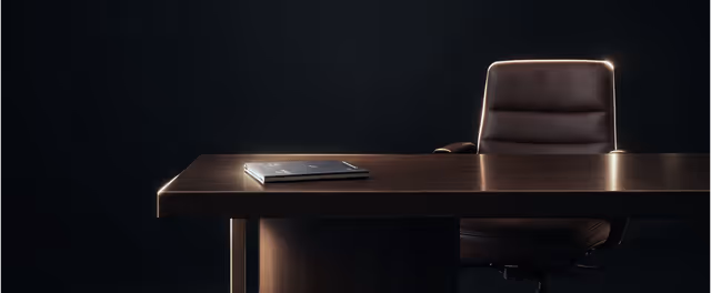 Dark office desk with a closed laptop and a black leather chair against a dark background.