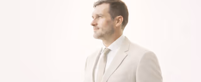 Man in light beige suit and tie looking to the side against a light background.