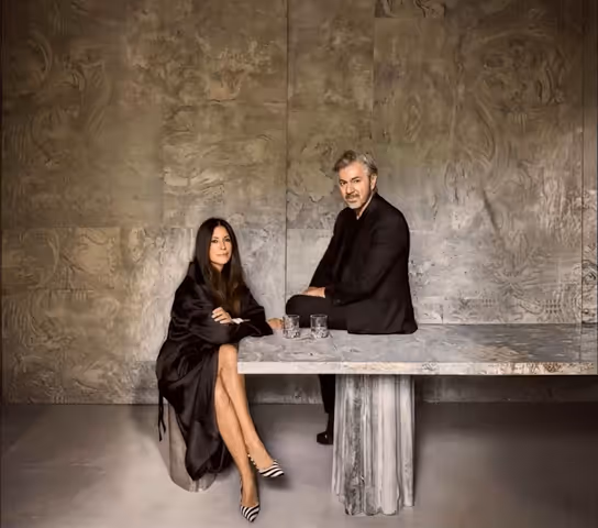 Woman in black dress sitting with crossed arms next to a man in dark suit seated on a marble table with two glasses in a modern, textured stone room.