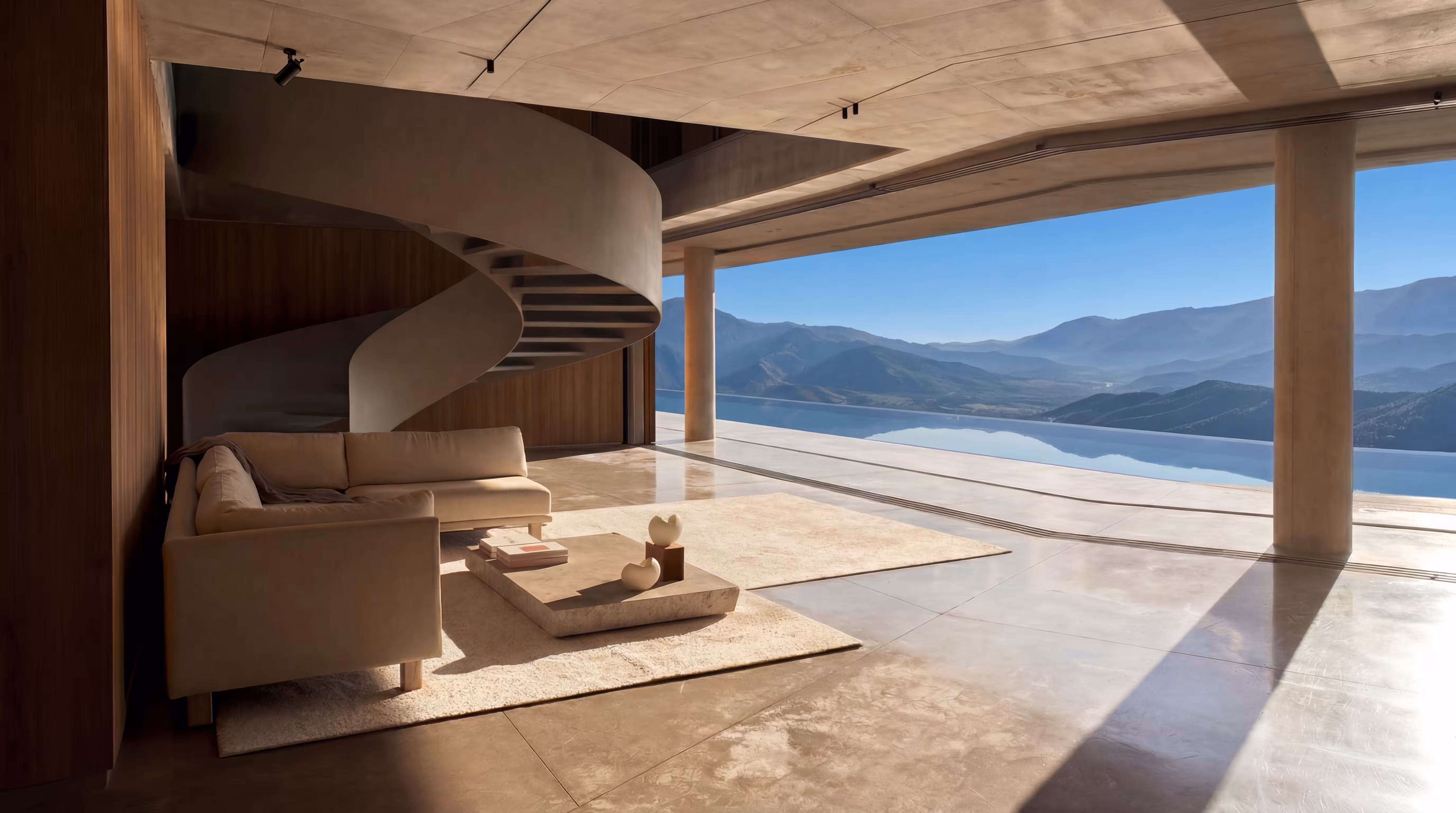 Modern living room with beige sofas, wooden chairs, concrete spiral staircase, and floor-to-ceiling windows overlooking a mountain and water view.