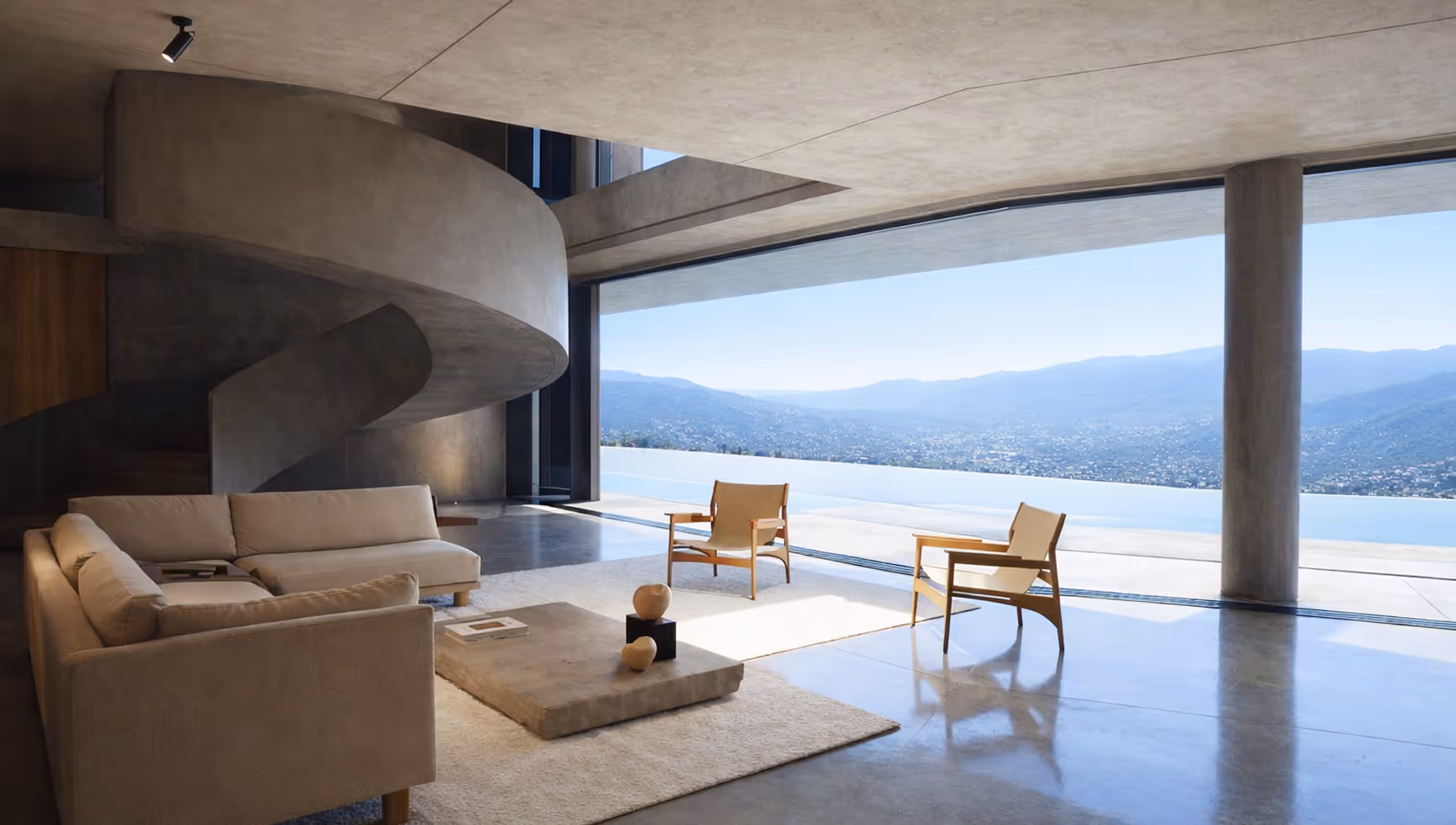 Modern living room with beige sofas, wooden chairs, concrete spiral staircase, and floor-to-ceiling windows overlooking a mountain and water view.