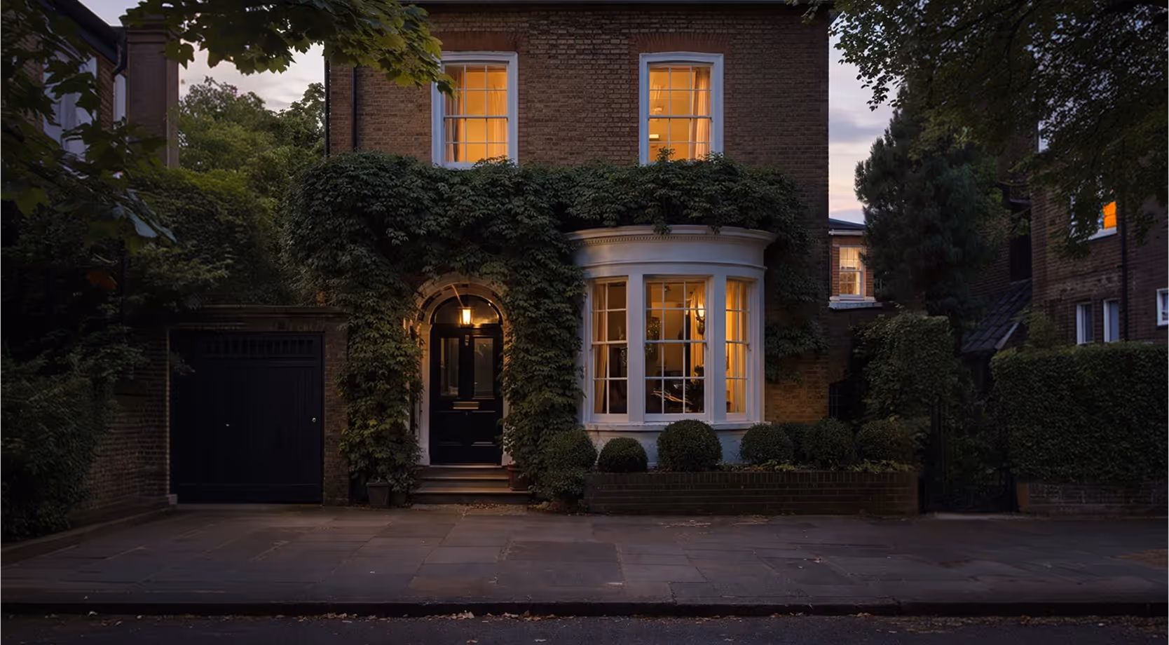 Two-storey brick house with a bay window and front door framed by ivy at dusk, with warm light glowing inside.