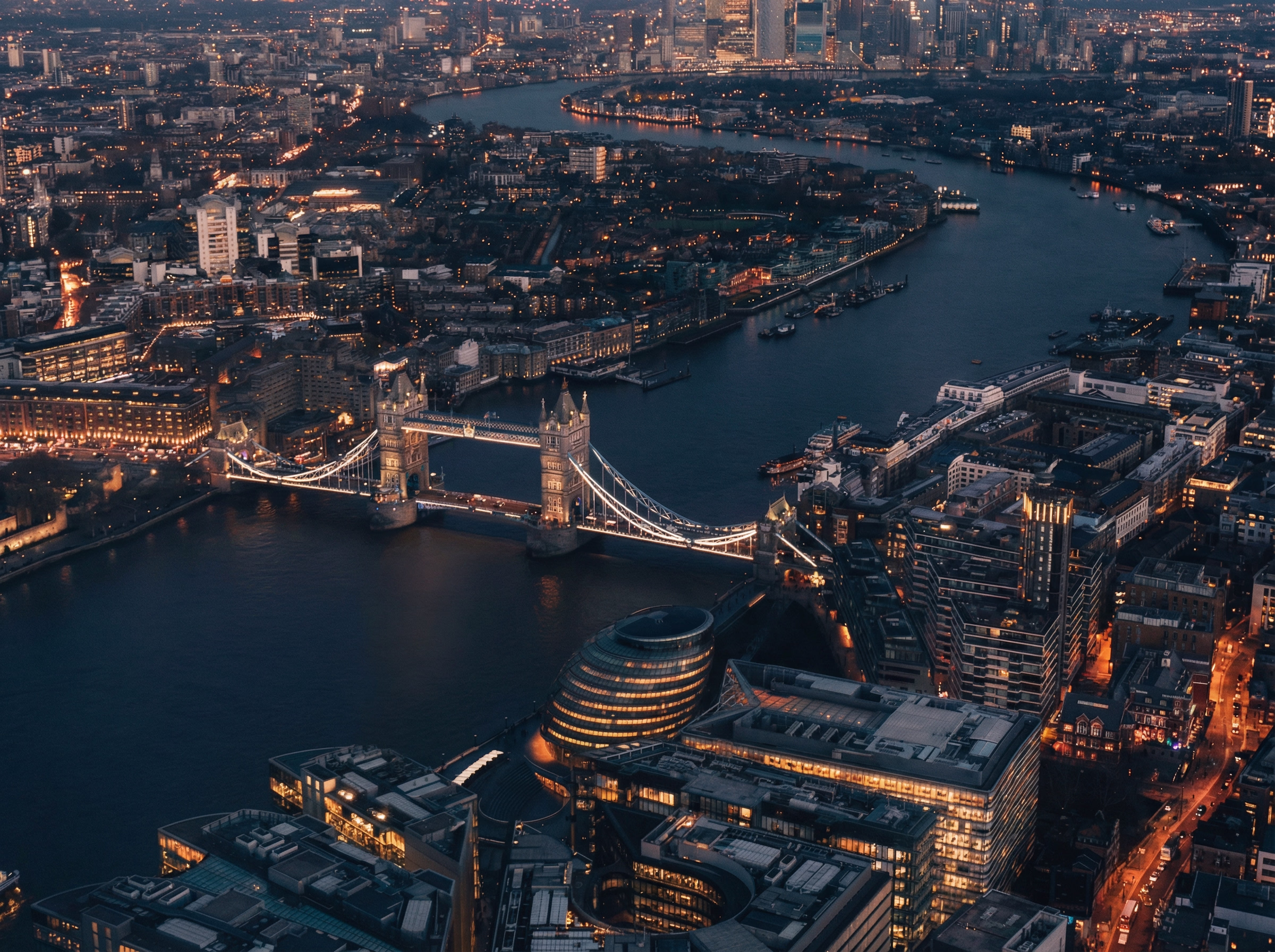 Aerial night view of Tower Bridge spanning the River Thames with surrounding illuminated buildings in London.