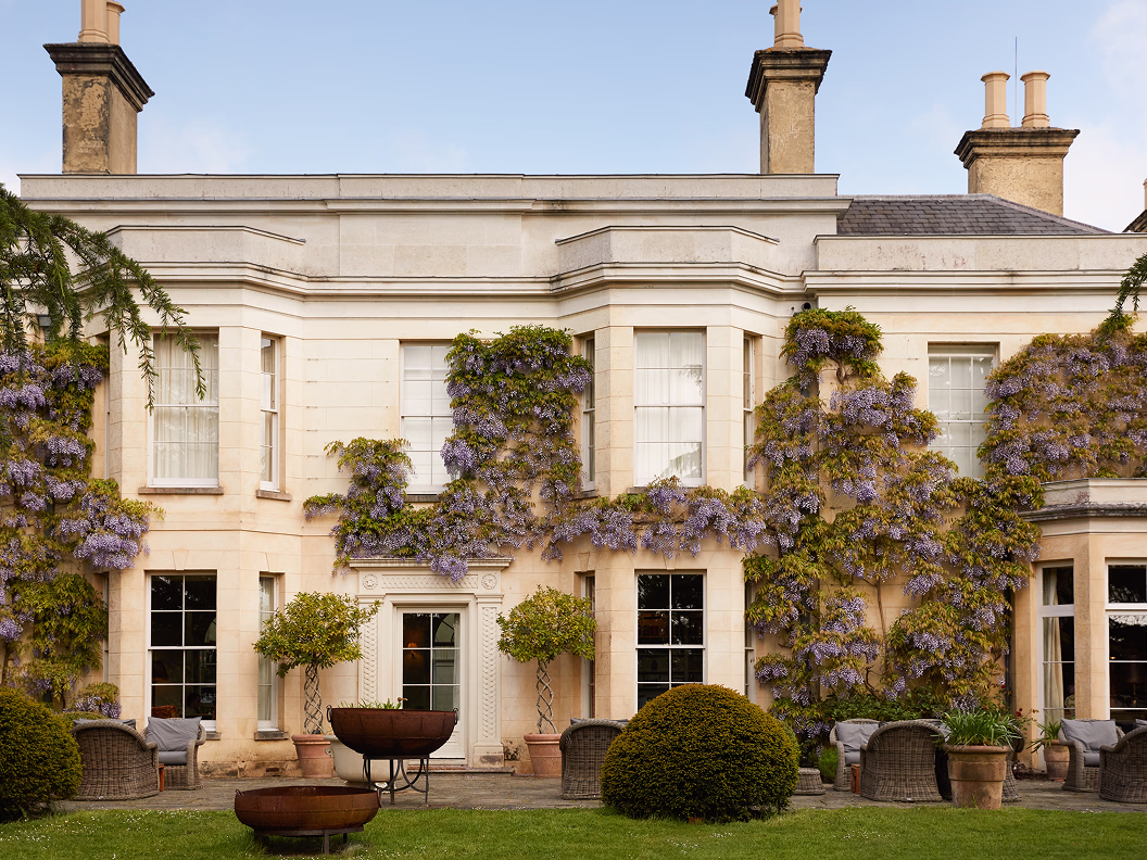 Elegant cream-coloured estate covered with lavender wisteria and surrounded by garden furniture and shrubs.