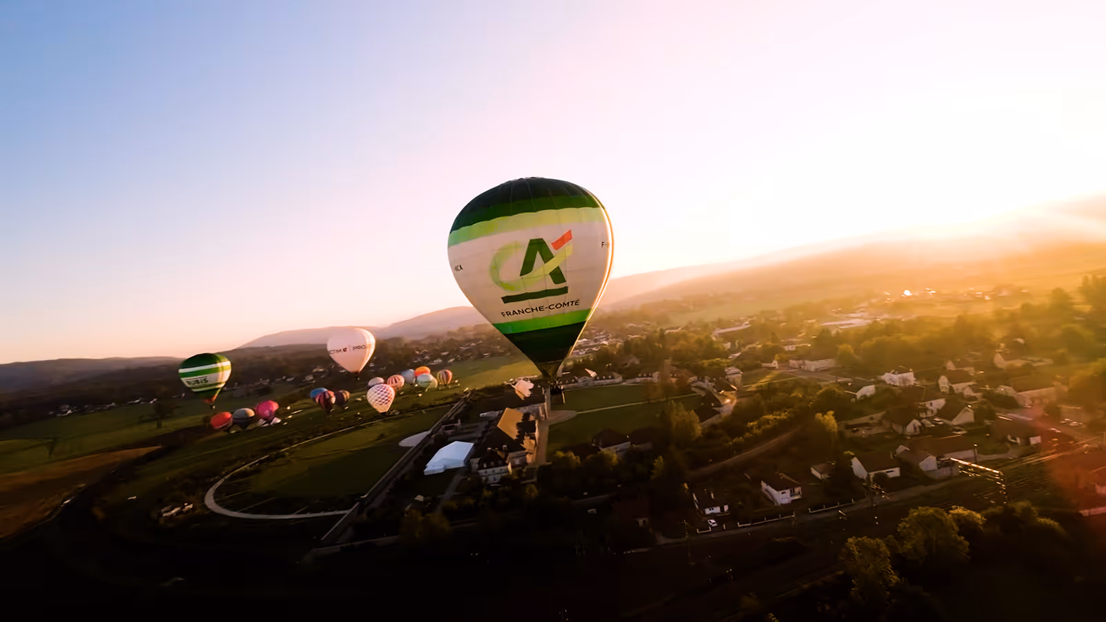 Montgolfière verte et blanche Crédit Agricole Franche-Comté en plein vol au-dessus d’un paysage rural au lever du soleil, avec plusieurs autres ballons colorés en arrière-plan.