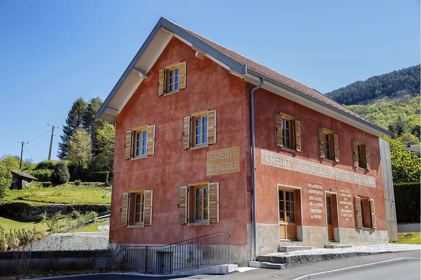 Façade d’une ancienne maison en pierre rouge abritant le Crédit Agricole Mutuel du Jura, située dans un environnement verdoyant et vallonné.