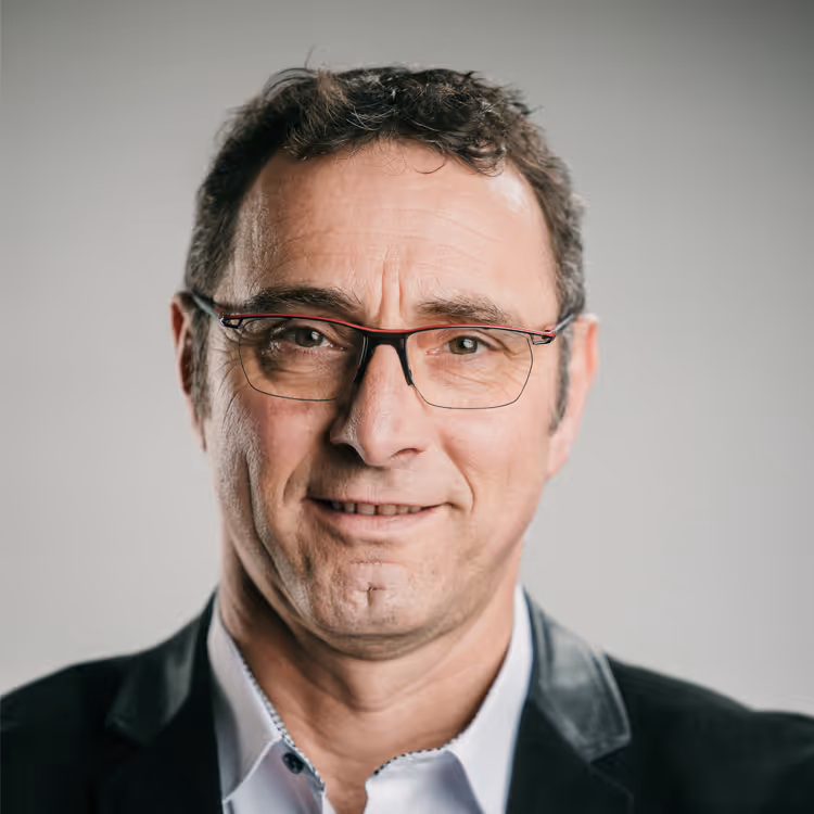 Portrait d’un homme en veste foncée et chemise claire, souriant légèrement, portant des lunettes rouges et noires, sur fond gris neutre.