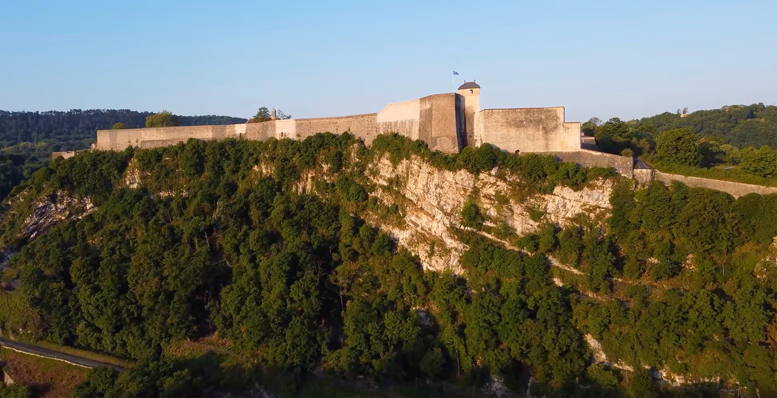Vue aérienne de la Citadelle de Besançon, fortification historique perchée sur une falaise boisée, au lever ou coucher du soleil.