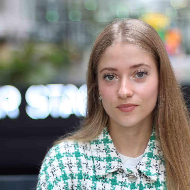 Portrait d’une jeune femme aux cheveux longs blonds, portant une veste texturée verte et blanche, posant avec un regard neutre dans un cadre flou en intérieur.
