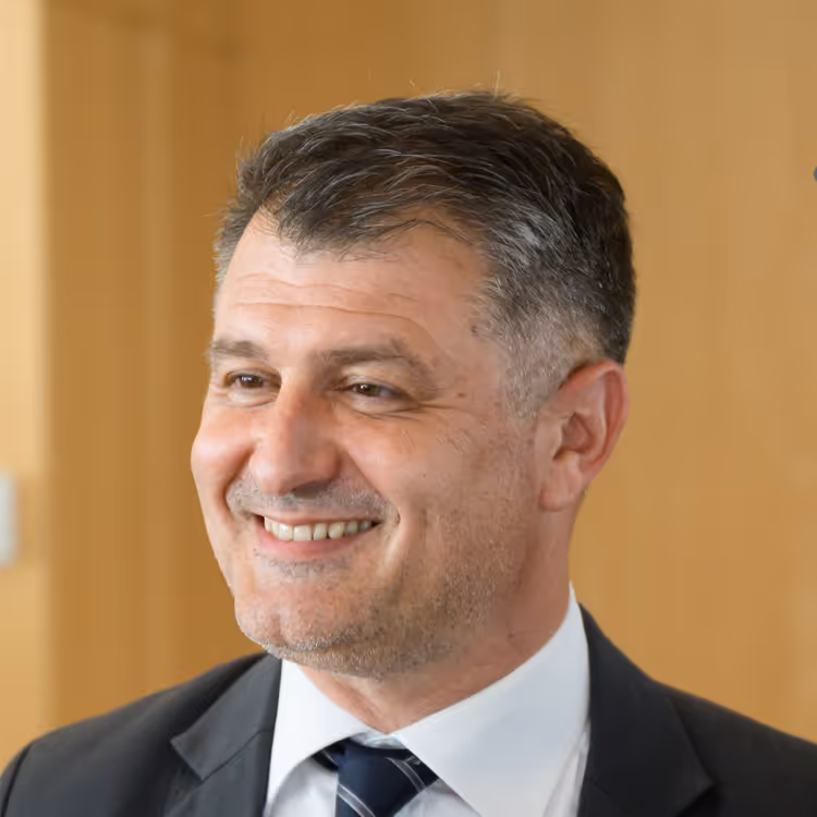 Portrait d’un homme en costume et cravate rayée, souriant dans un environnement professionnel lumineux en bois clair.