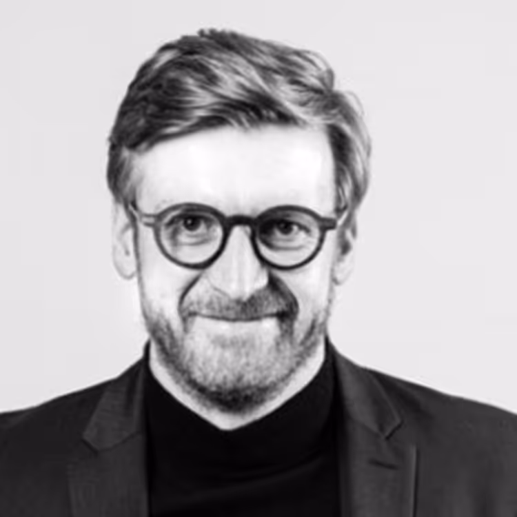 Portrait en noir et blanc d’un homme souriant, portant des lunettes rondes et un col roulé noir, sur fond clair.