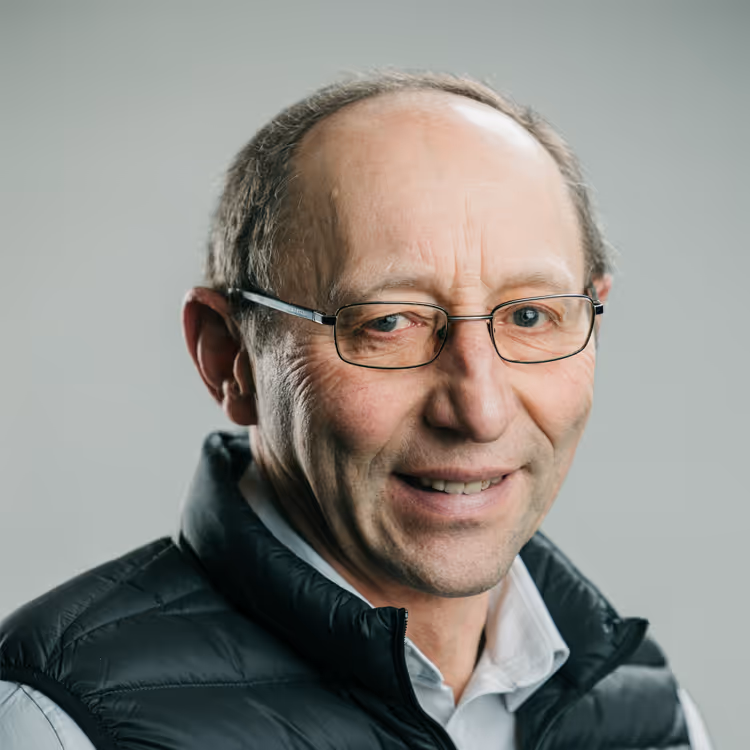 Portrait d’un homme portant des lunettes et une doudoune noire, avec chemise blanche, souriant sur fond gris clair.