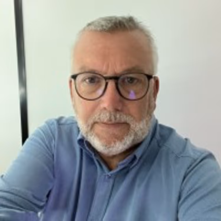 Portrait d’un homme barbu aux cheveux gris, en chemise bleue et lunettes noires, photographié en intérieur devant un mur blanc.