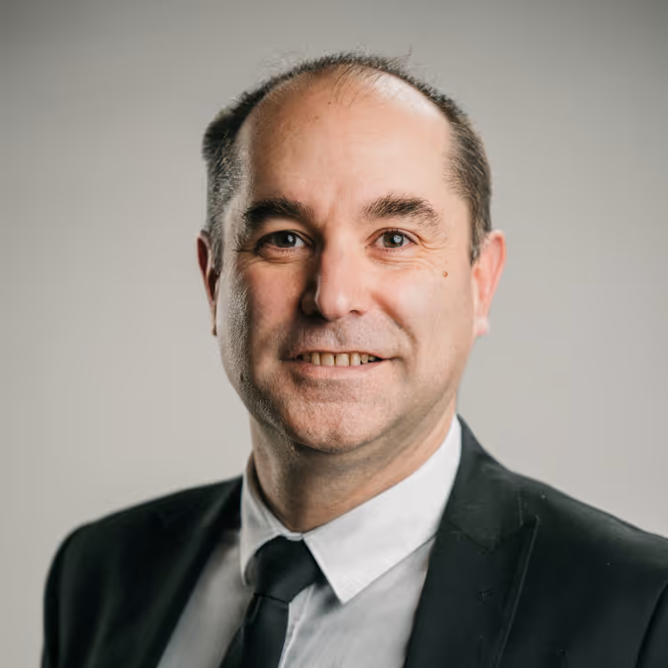 Portrait d’un homme en costume noir, chemise blanche et cravate, souriant légèrement sur fond gris neutre.