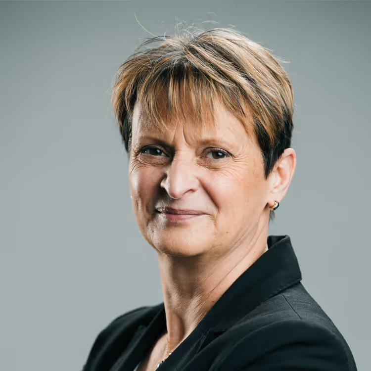 Portrait d’une femme mûre en veste noire, cheveux courts avec mèches claires, sourire discret sur fond gris.