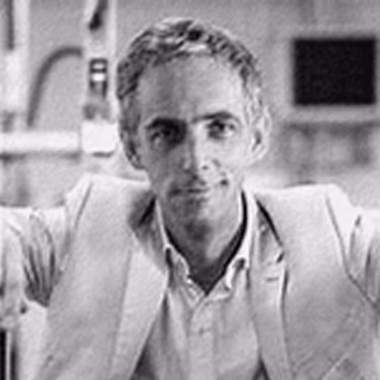 Portrait en noir et blanc d’un homme en veste claire, regard direct et expression posée, photographié dans un cadre professionnel flou.