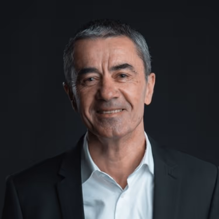 Portrait d’un homme en costume sombre et chemise claire, souriant légèrement, photographié sur fond noir.