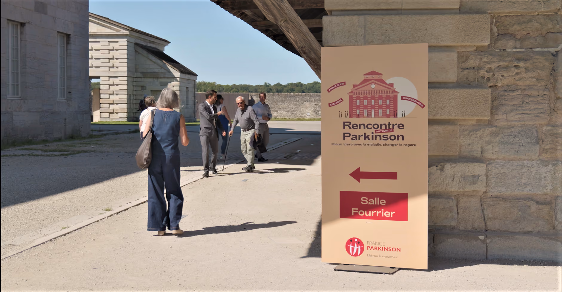 Au premier plan, un panneau indiquant l'événement "Rencontre avec Parkison" et en arrière-plan, trois personnes se dirigeant vers l'événement