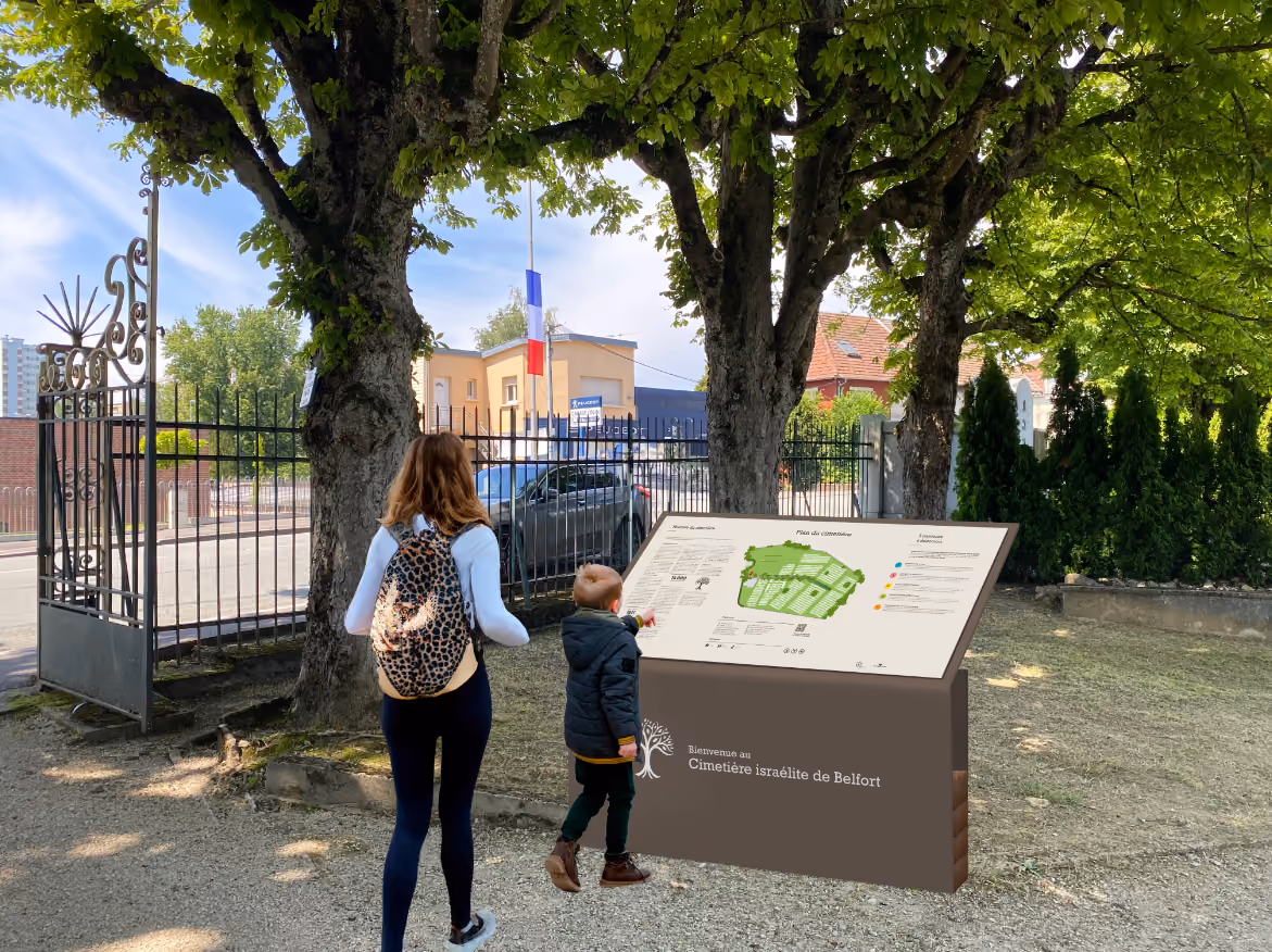 image de l'entrée du cimetière israélite de Belfort. Un panneau avec dessus le plan du cimetière est visible, avec au premier plan une femme et un enfant qui le regarde