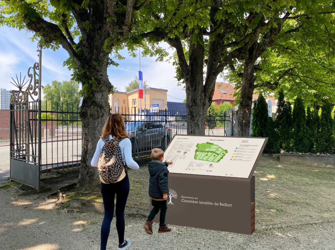 image de l'entrée du cimetière israélite de Belfort. Un panneau avec dessus le plan du cimetière est visible, avec au premier plan une femme et un enfant qui le regarde
