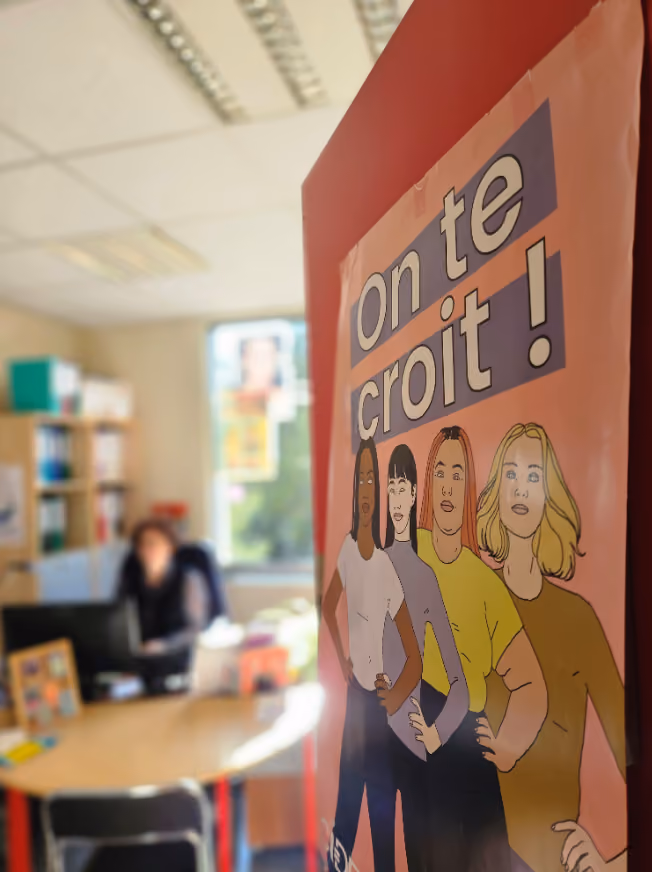 Au premier plan, une porte ouverte avec un poster sur lequel on voit des femmes dessinées avec marqué "on te croit !" donnant sur un bureau en arrière-plan floutté avec une femme a son bureau devant son ordinateur