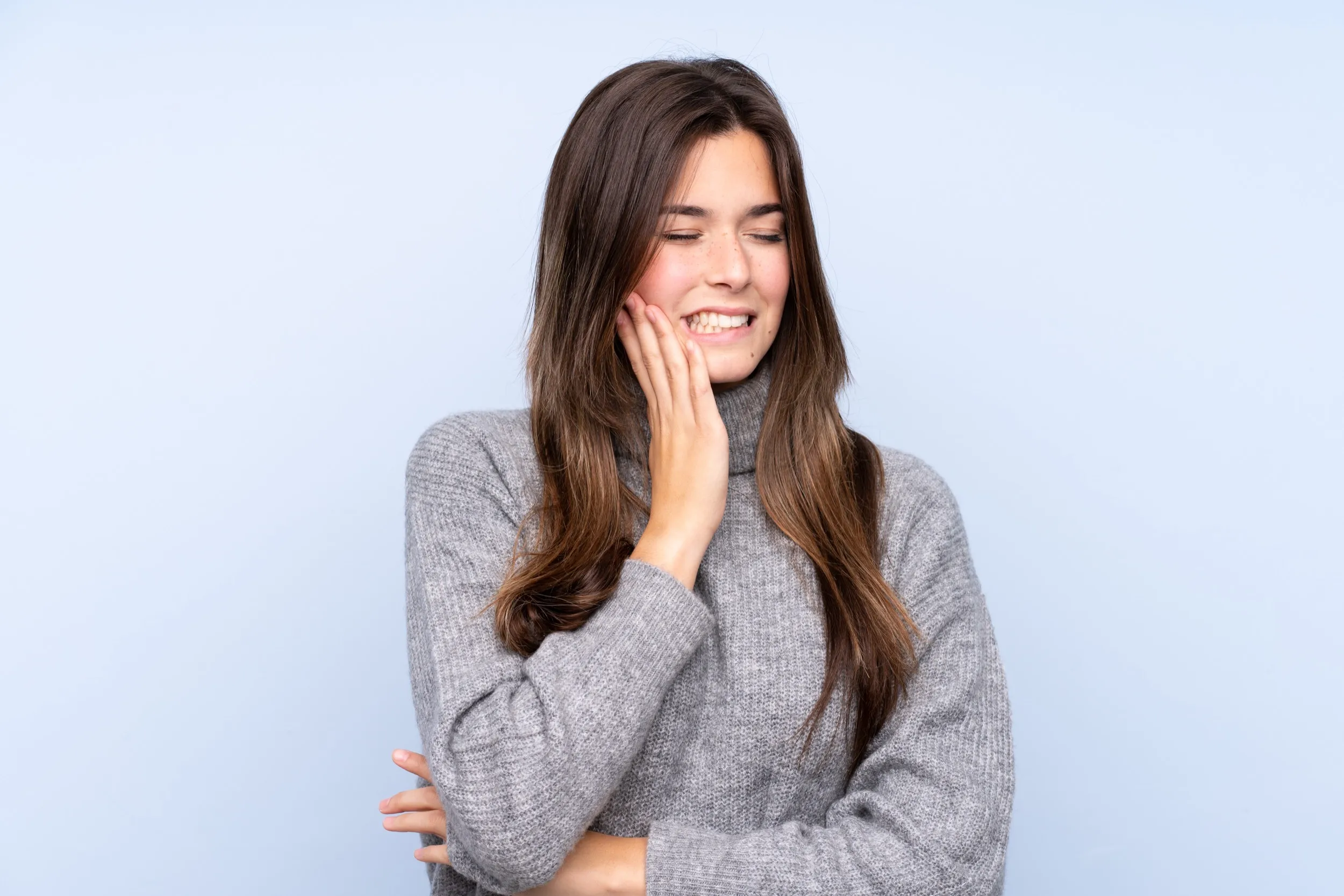 Woman in grey sweater smiling with hand on cheek against blue background