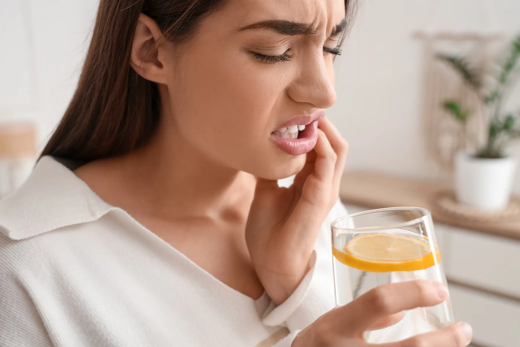 Woman wincing in pain, holding cheek while drinking lemon water
