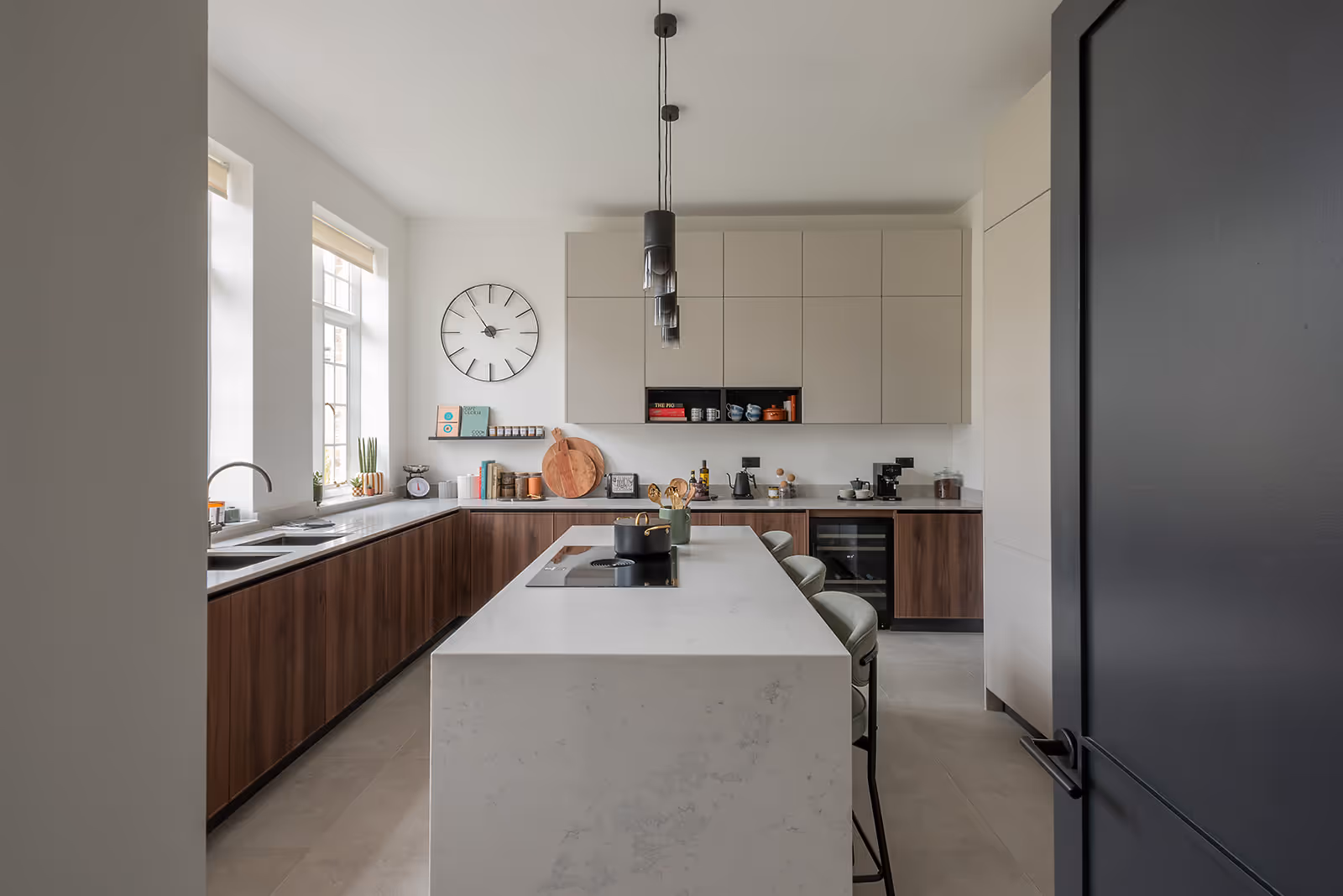 Modern kitchen with cream upper cabinets and walnut lower units, featuring a white marble island with bar stools, polished concrete flooring, and pendant lighting. Large windows provide natural light to the neutral-toned space.