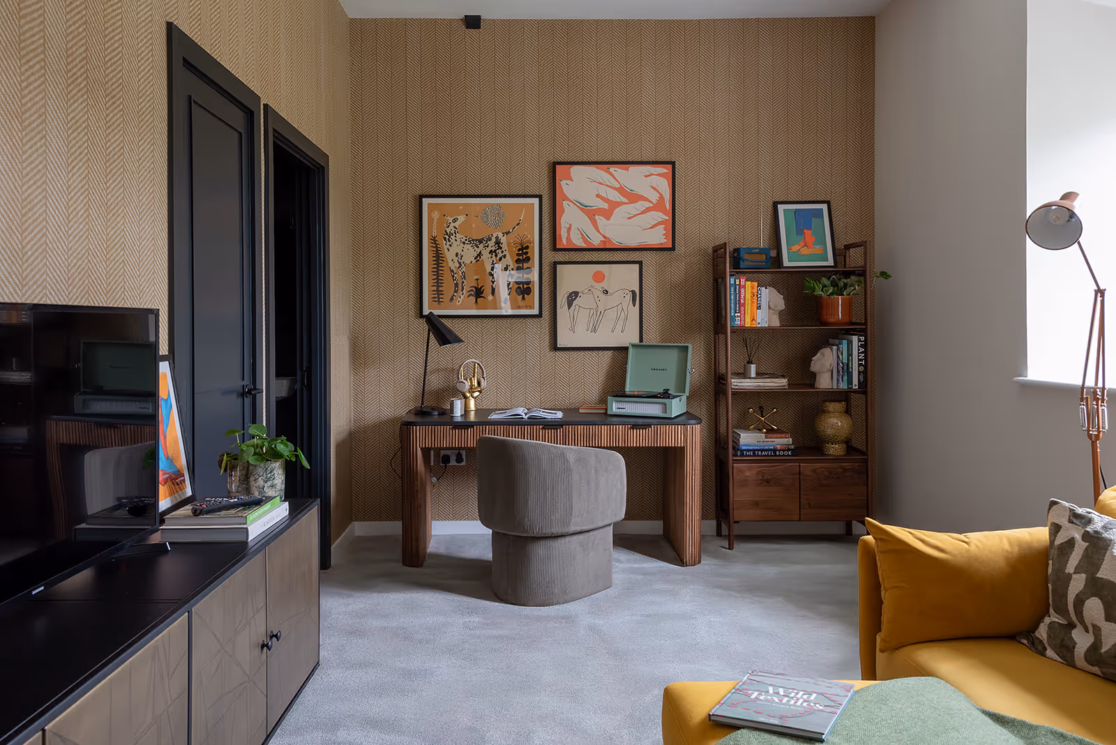 Home office space with herringbone patterned wallpaper, walnut desk and shelving unit, grey upholstered chair, curated gallery wall of contemporary art, and mustard yellow sofa visible in foreground on polished concrete flooring.