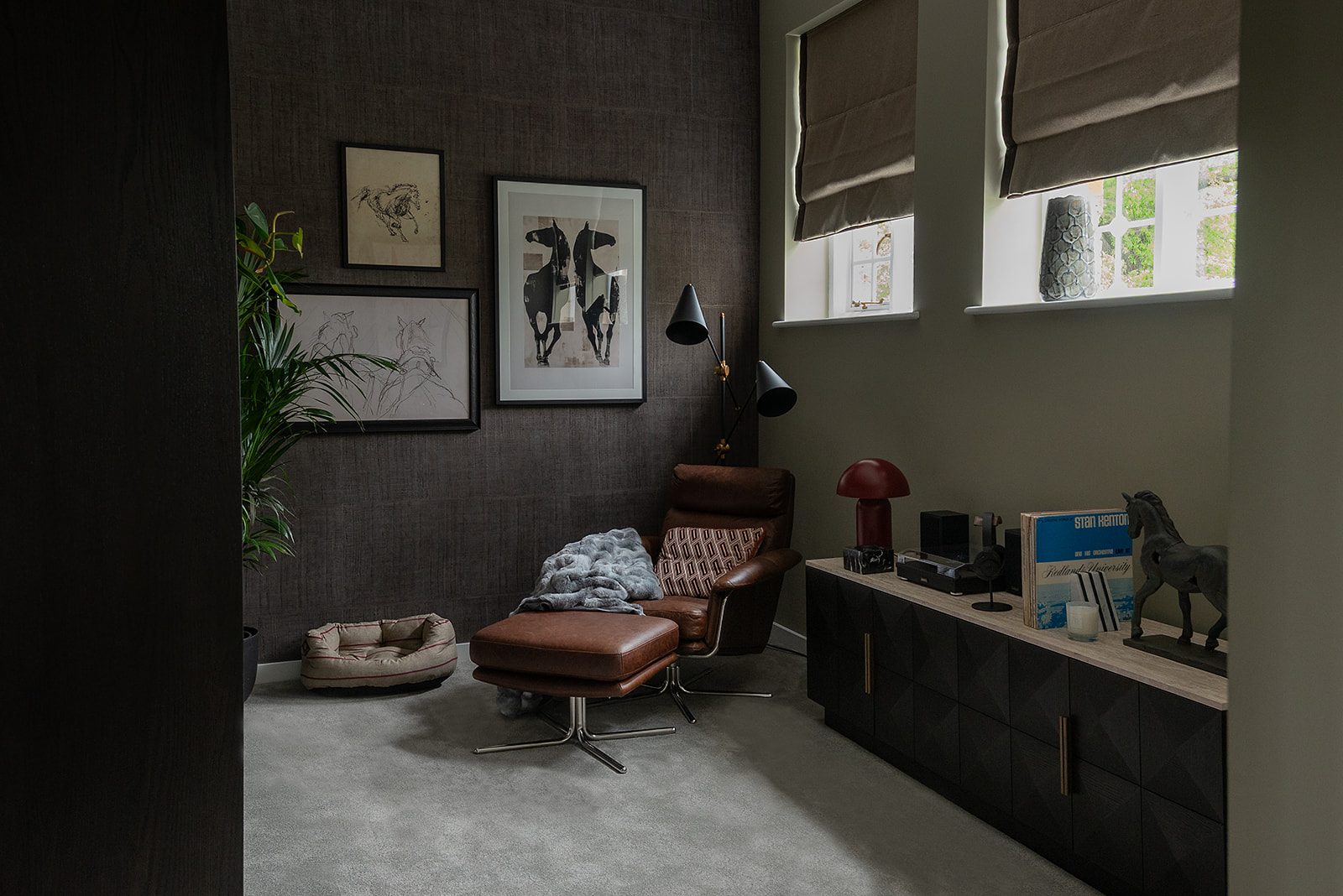 Cosy reading corner with a cognac leather Eames-style chair and ottoman, dark textured wallpaper, gallery wall of framed botanical prints, built-in shelving with books, and a mushroom-shaped table lamp on polished concrete flooring.