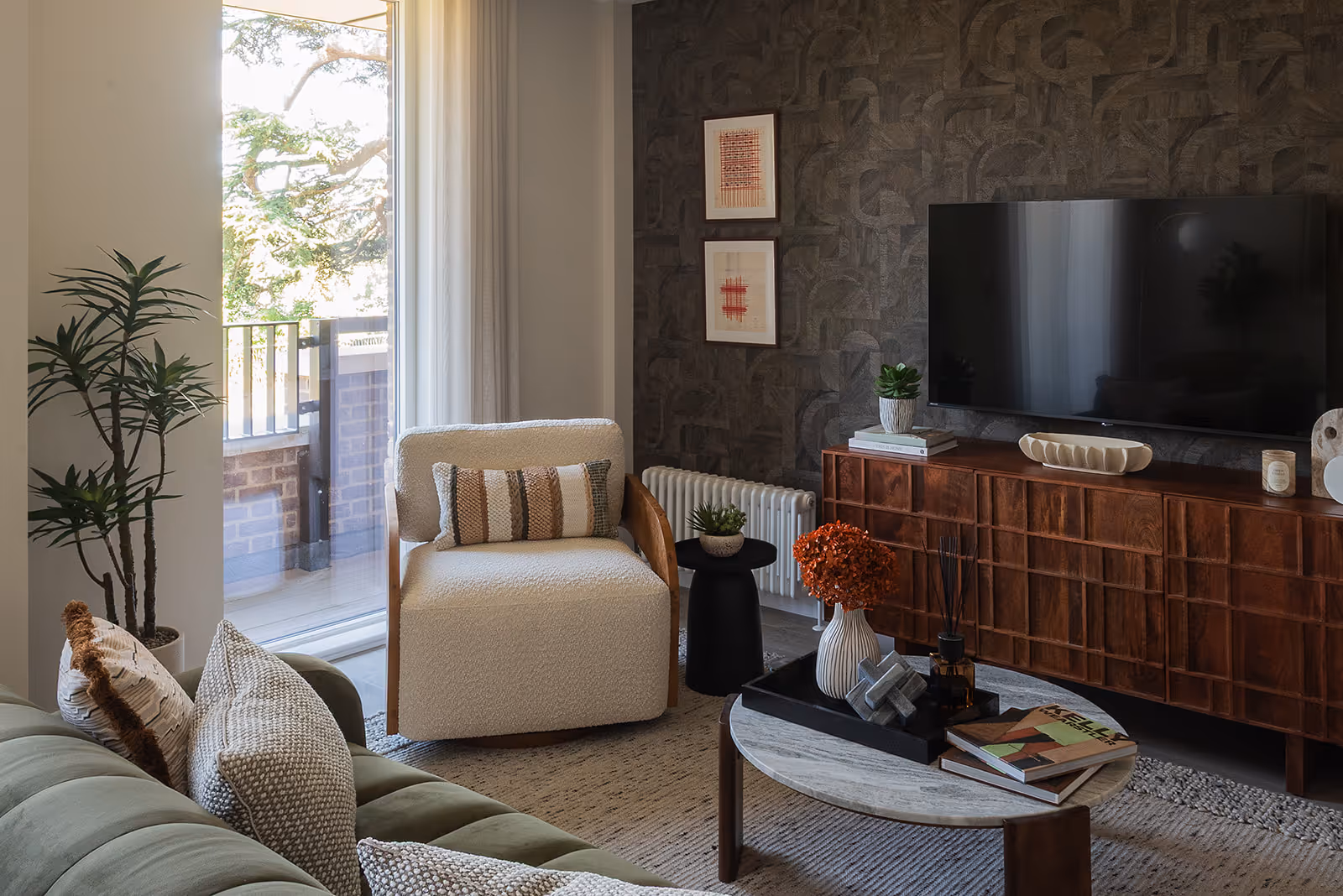 A modern living room featuring a textured stone accent wall, cream armchair with striped cushions, olive green sofa with knitted throw pillows, and a walnut media unit beneath a wall-mounted television. A round coffee table sits atop a jute rug, with large windows revealing a leafy outdoor view.