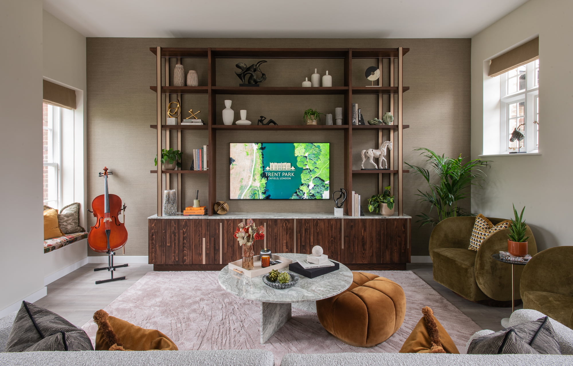 Elegant modern living room interior featuring a wooden wall unit with decorative shelving, flat-screen TV displaying “Trent Park, Enfield, London,” and stylish furniture including green armchairs, marble coffee table, cello, and window seat. Natural light, neutral tones, and curated decor create a sophisticated, contemporary home aesthetic.