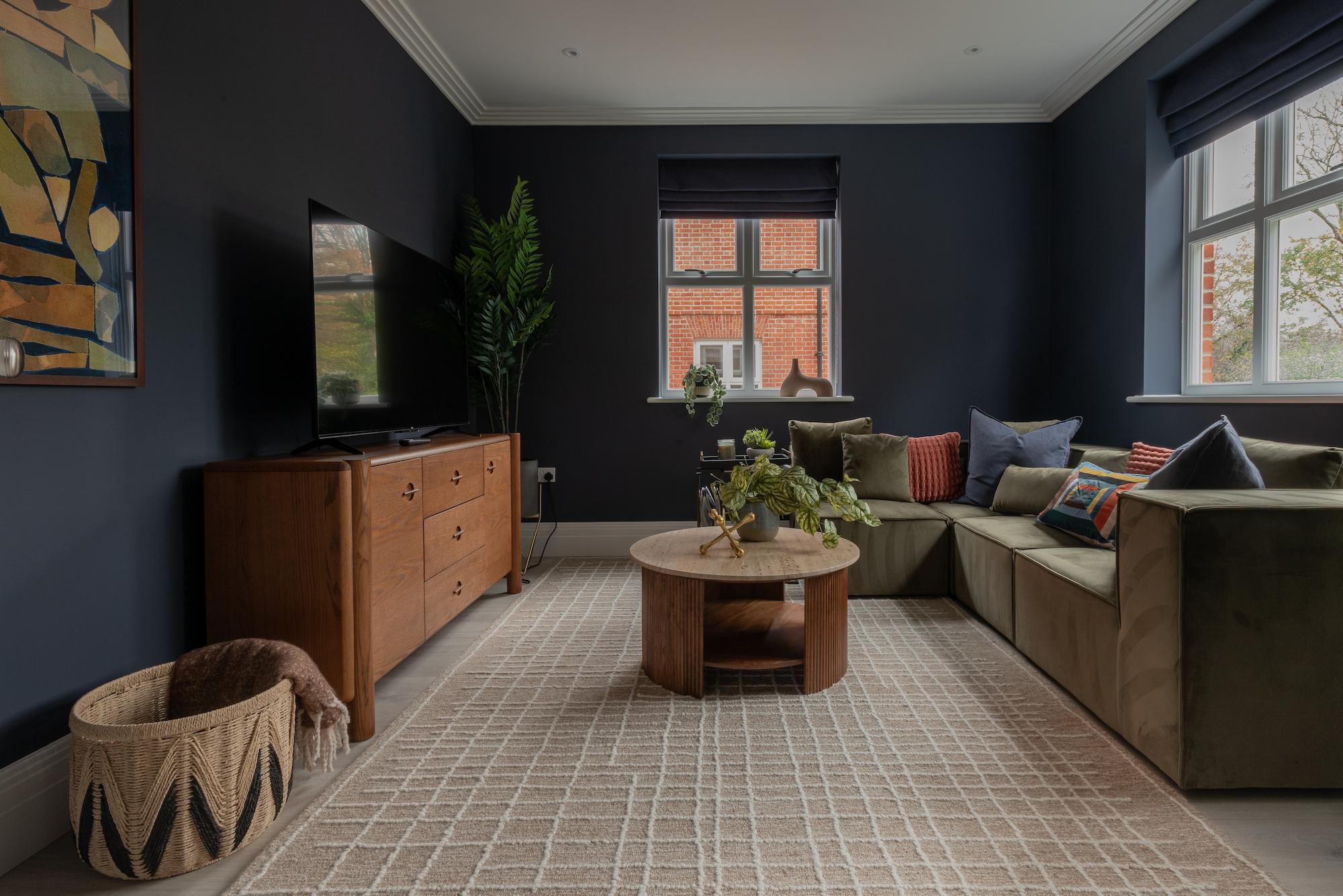 Modern living room with dark charcoal walls, beige L-shaped sectional sofa with colourful patterned cushions, round wooden coffee table with plants, mid-century modern wood credenza with wall-mounted TV, tall artificial palm plant, and woven basket in foreground. Large windows with dark roman blinds show brick buildings outside.