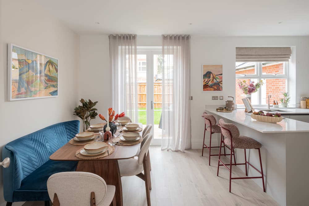 Open-plan dining area and kitchen with white walls and pale wood flooring. Royal blue velvet banquette seating along oval wood dining table set with beige plates and orange floral centrepiece. White sheer curtains frame French doors leading to garden. Kitchen features white worktops, woven bar stools, and abstract artwork on walls.