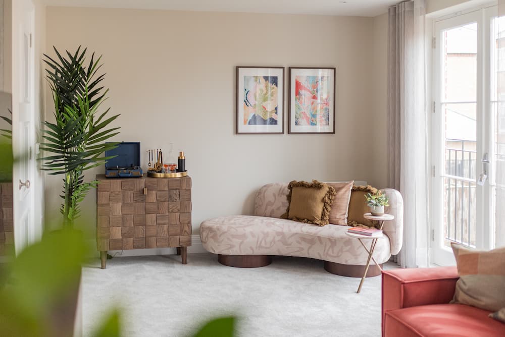 Bright living room with cream walls featuring curved chaise lounge upholstered in leaf-patterned fabric with textured tan cushions. Wooden chest of drawers with basket-weave pattern supports TV and decorative items. Two abstract art prints in wooden frames hang above. Tall artificial palm plant stands to left. French doors with sheer curtains open to balcony. Coral ottoman visible in corner.