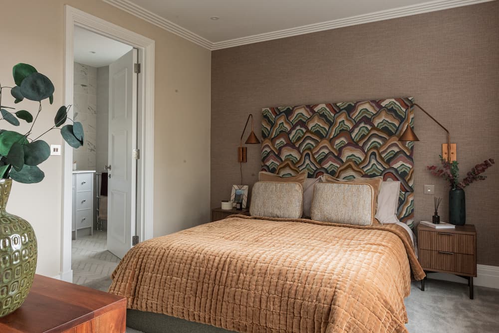 Contemporary bedroom with textured taupe feature wall behind bed. Colourful art deco-style upholstered headboard in geometric wave pattern with earth tones and teal. Rust-coloured quilted bedding with cream pillows. Mid-century wooden nightstands with grooved fronts flank bed, topped with copper wall sconces. White crown moulding frames ceiling. Doorway leads to marble-tiled ensuite bathroom. Large decorative vase with artificial eucalyptus in foreground.