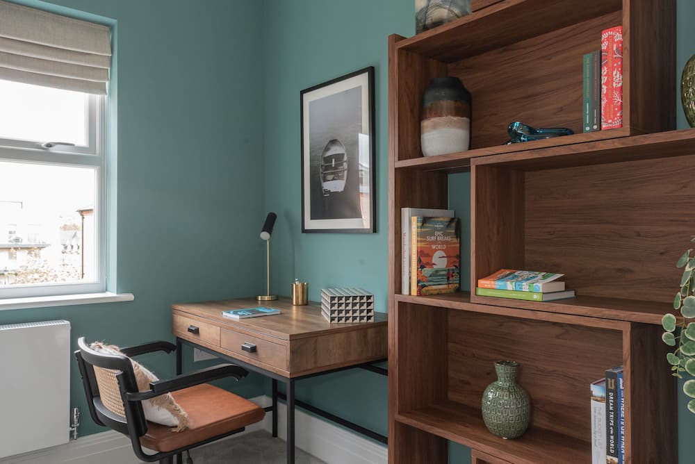 Home office with teal-painted walls featuring mid-century modern wooden desk with two drawers on metal frame. Black framed surfboard photograph hangs above desk alongside small desk lamp and geometric box. Wooden modular shelving unit displays books, decorative vases including green textured piece, and red patterned book standing upright on top shelf. Black chair with terracotta cushion sits at desk. Artificial eucalyptus visible on right edge. Window with roman blind on left shows rooftops outside.