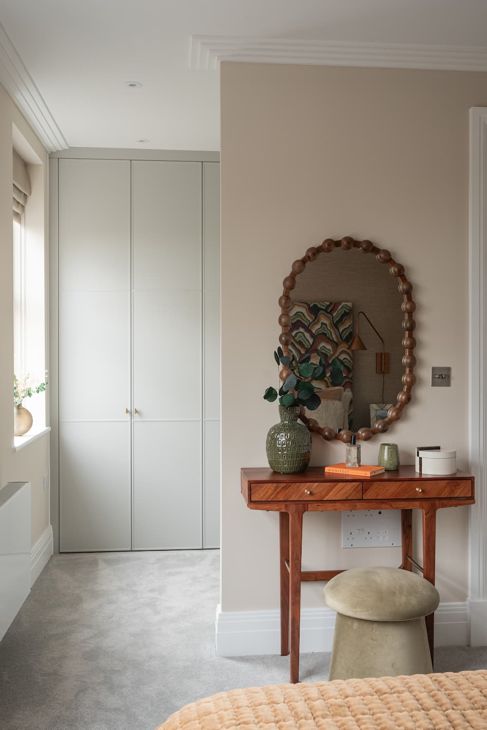 Bedroom dressing area with built-in white floor-to-ceiling wardrobes on left and pale taupe feature wall on right. Wooden console table with two drawers holds decorative vase with artificial eucalyptus, small textured vase, orange book, and candle. Large circular mirror with decorative scalloped copper frame hangs above. Cream upholstered mushroom-style stool sits underneath. White electrical sockets visible on wall. Rust-coloured quilted bedding visible in foreground.