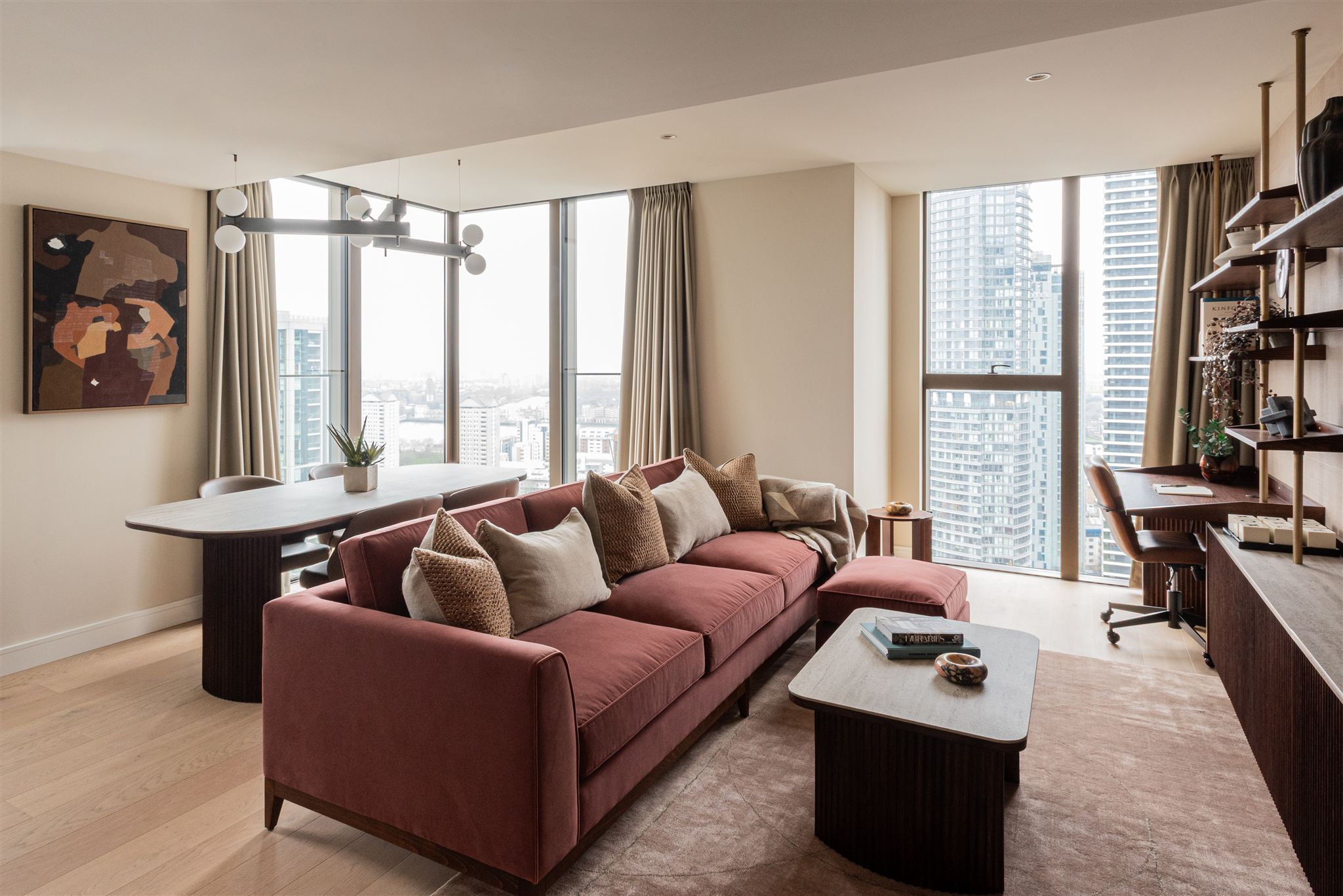 Contemporary apartment living space with terracotta three-seater sofa adorned with textured cushions, overlooking panoramic city views through floor-to-ceiling windows. The room includes a marble-top coffee table, walnut shelving system, and abstract artwork in warm tones. High-rise buildings visible through multiple window panels.