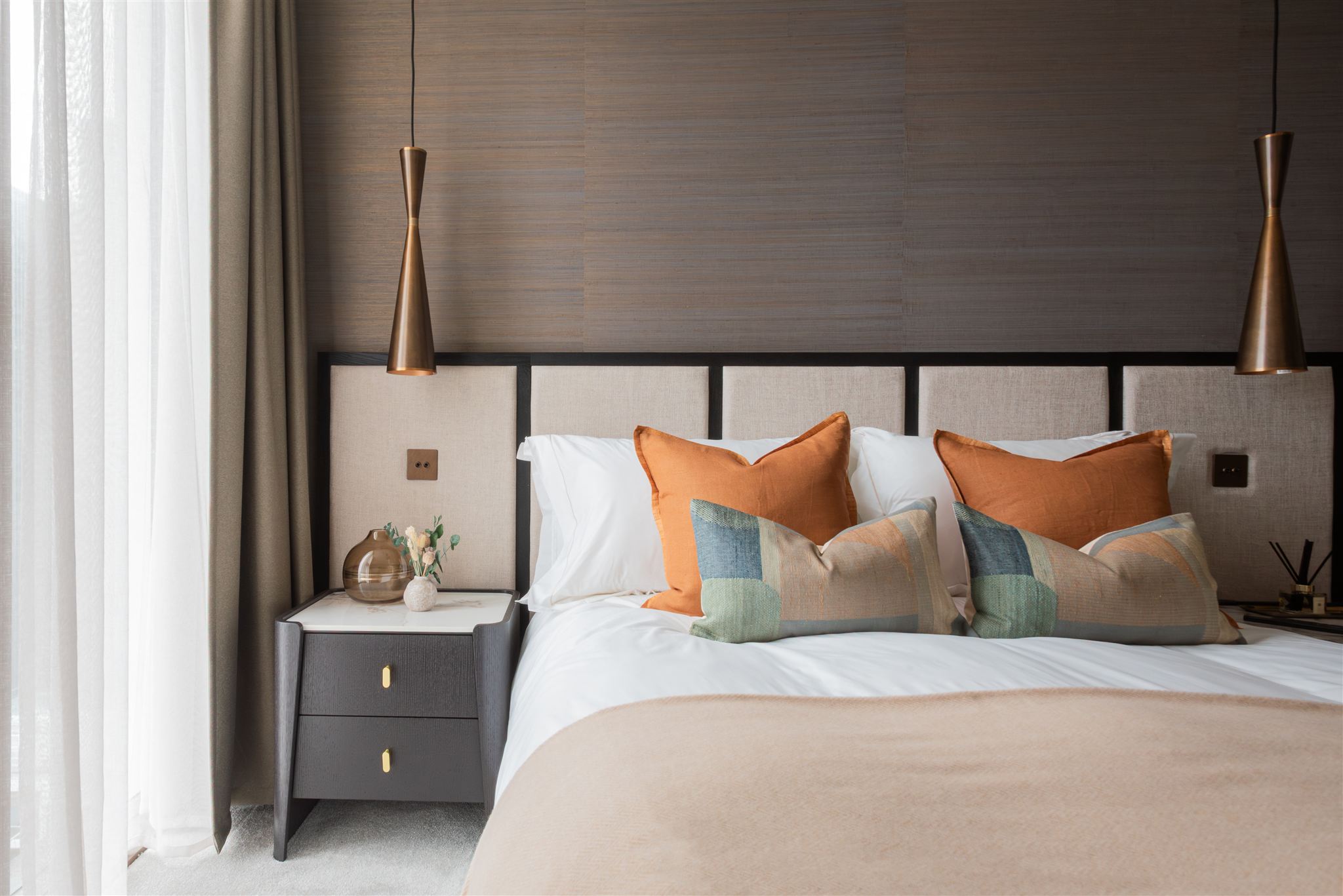 Luxurious bedroom with upholstered headboard featuring vertical panelling in cream linen, dressed with white bedding, burnt orange cushions, and blue-green striped accent pillows. Brass conical pendant lights hang either side of the bed, with grey bedside tables featuring gold handles.