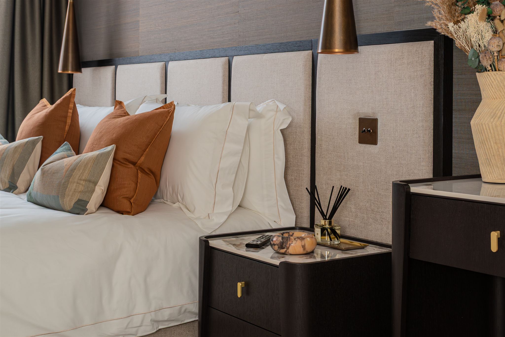 Close-up of hotel-style bedroom styling showing layered pillows in burnt orange linen and colour-blocked blue-green cushions against white bedding. A dark wood bedside table with brass handles displays a reed diffuser and decorative bowl, with dried flowers in a textured vase visible to the right.