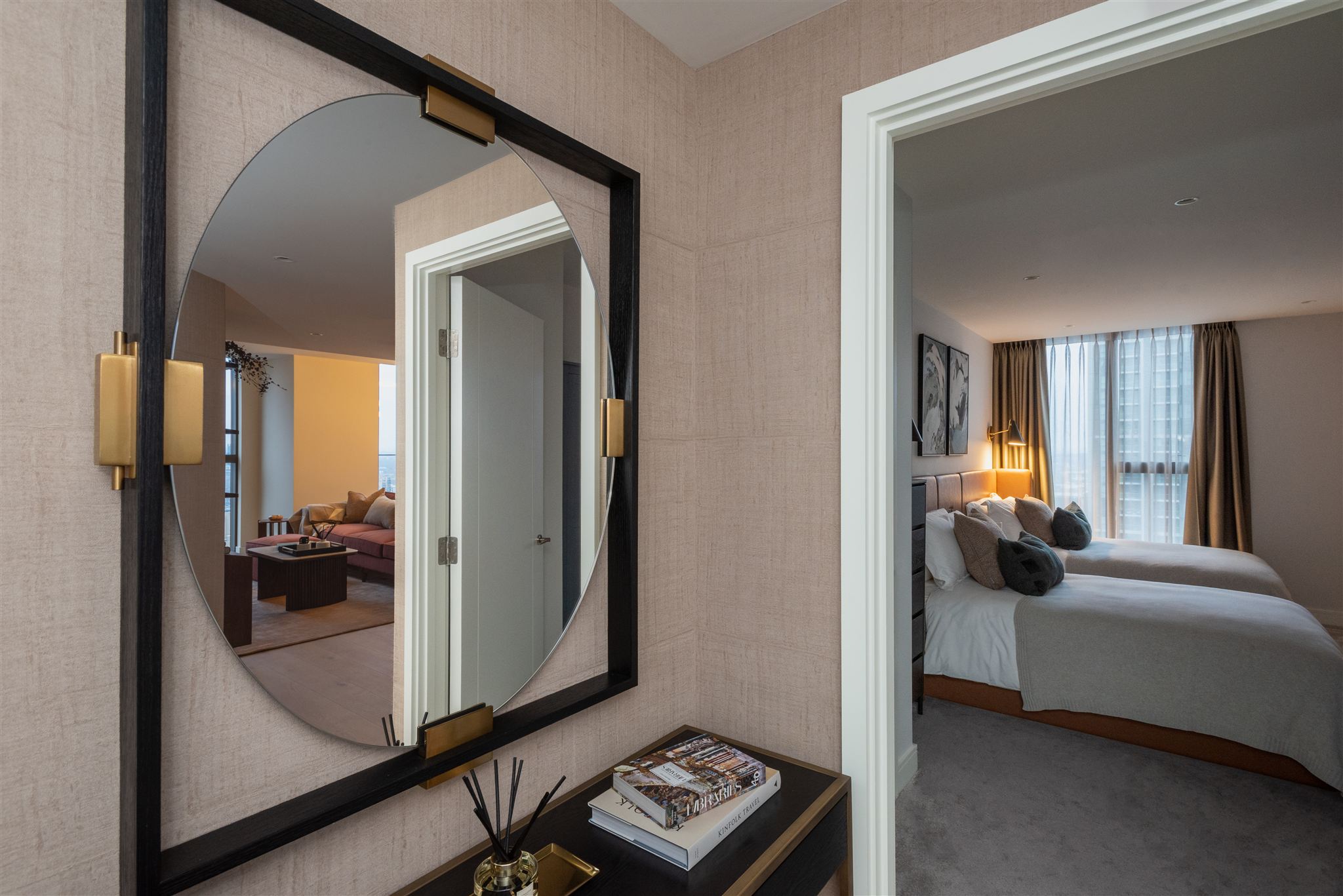 Hallway perspective showing the same decorative mirror and console arrangement, with views through white-painted doorways into adjacent living and bedroom spaces. The mirror reflects warm evening lighting from the living area, whilst a grey-toned bedroom is visible through the right doorway.