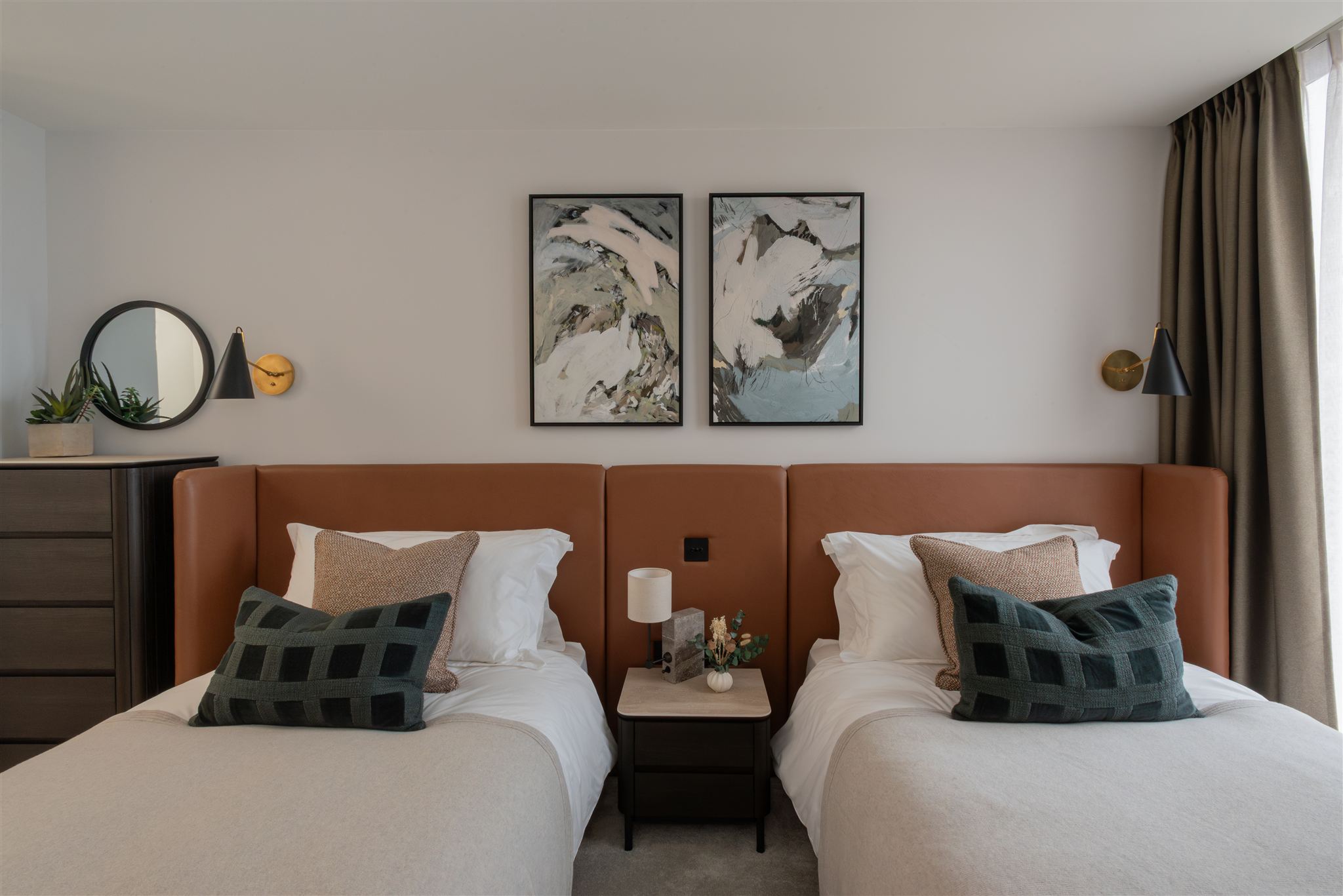 Twin bedroom with tan leather upholstered headboard spanning both beds, dressed with white linens and layered cushions in textured beige and dark green check patterns. Two framed abstract mountain artworks in muted tones hang above, with black conical wall lights and a small shared bedside table between the beds.
